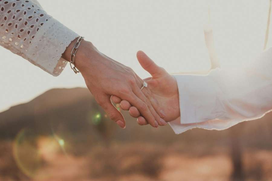 Gros plan intime sur les mains enlacées d'un couple dans le désert au coucher du soleil, avec une bague de fiançailles bien visible au doigt de la femme.
