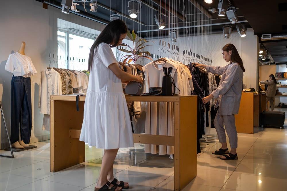 Deux femmes dans une boutique de mode, l'une en robe blanche et l'autre en ensemble gris, cherchent à bien s’habiller.