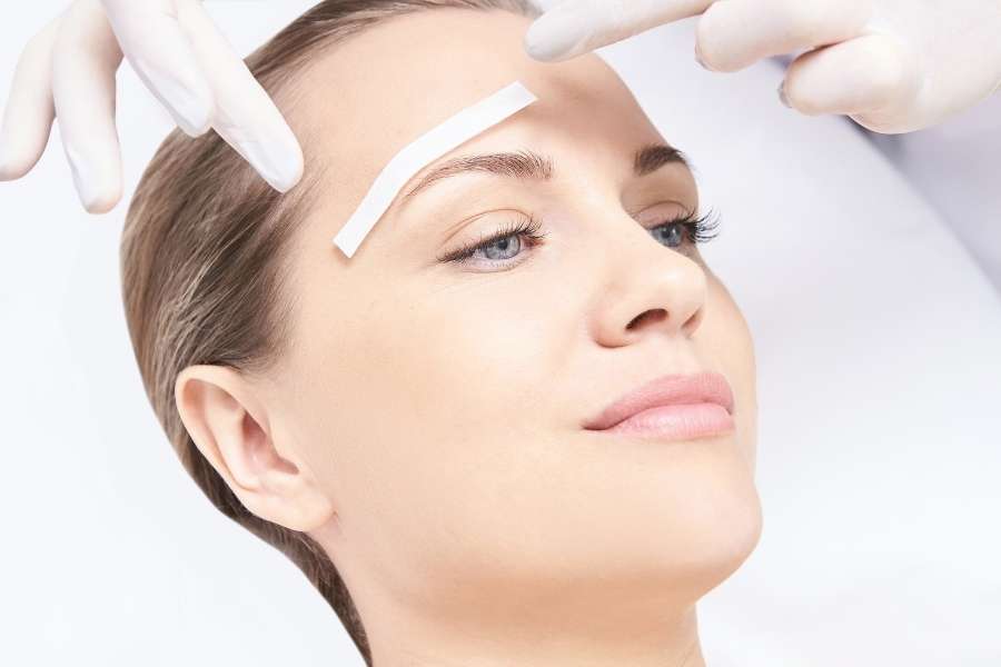 Une esthéticienne en gants blancs mesure et positionne un guide adhésif pour redessiner des sourcils sur une cliente.