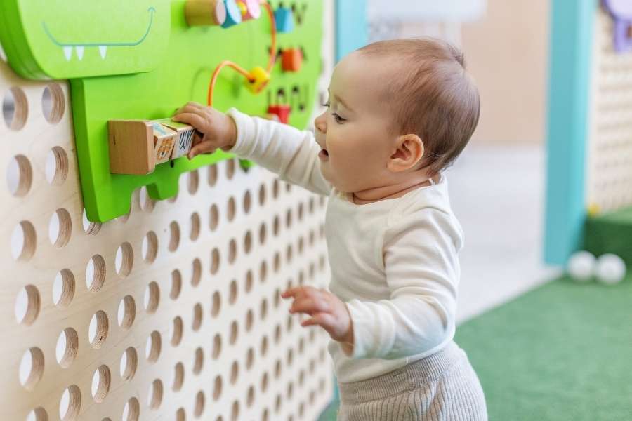 Un bébé en tenue claire debout et concentré devant un grand panneau mural vert avec des trous, explorant des activités d'éveil sensoriel.