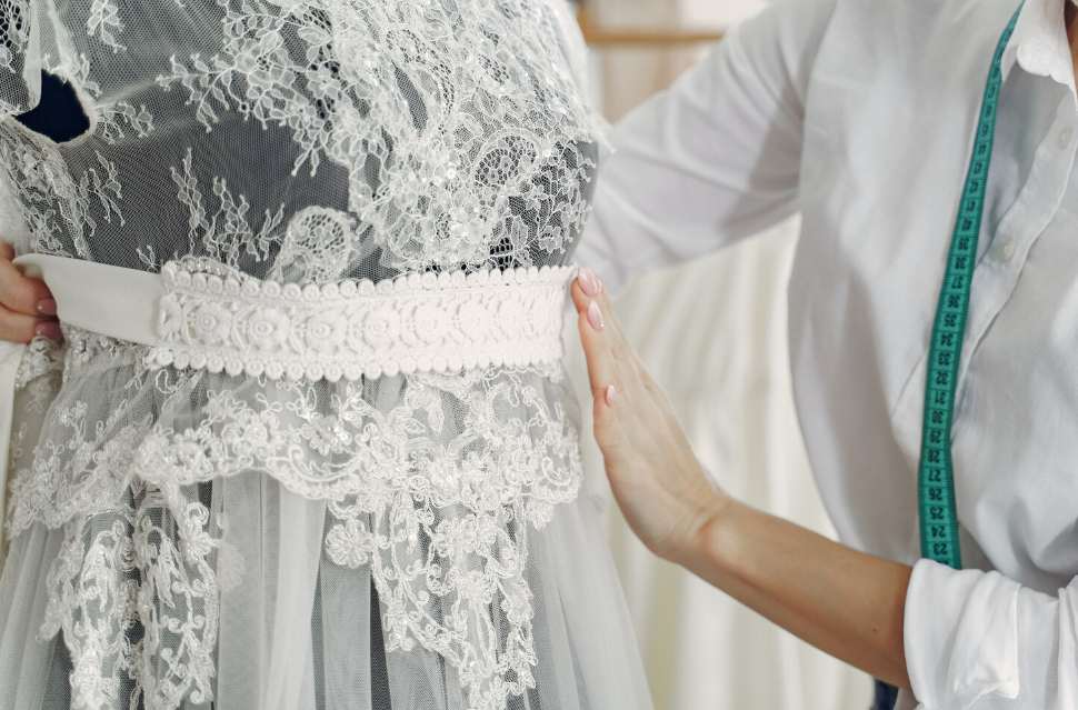 Une couturière avec un mètre ruban ajuste la ceinture en dentelle d'un modèle blanc pour savoir où acheter une robe de mariage d'occasion et la retoucher.